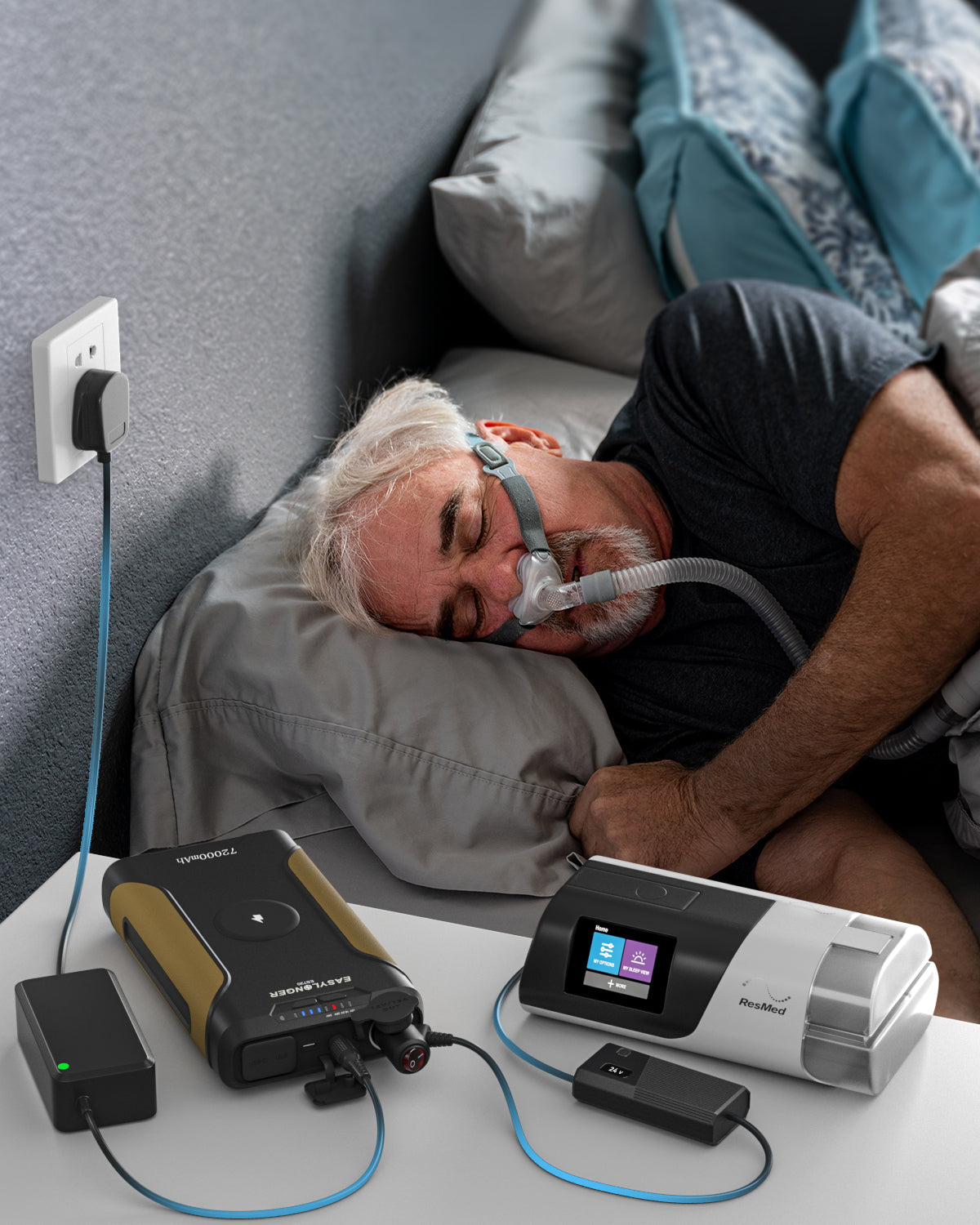 An older man lies in bed with a CPAP mask, next to the EASYLONGER Universal CPAP DC Power Cord (12V/24V) car charger for ResMed & Philips on the bedside table; pillows and bedding are visible, framed in 16:9.