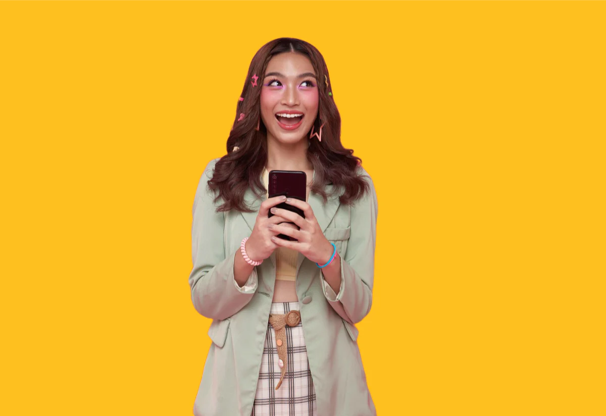 A young woman with long brown hair holds a smartphone and smiles excitedly. She is wearing a light green jacket over a plaid outfit, with pink hair clips, standing against a solid yellow background.