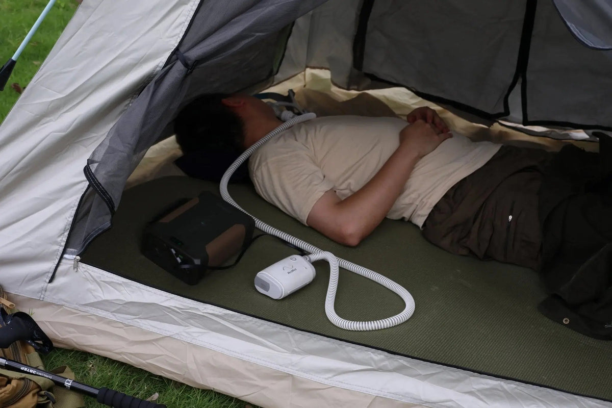 Person wearing a CPAP mask in a tent, with tubing, a compact CPAP machine and an EASYLONGER battery