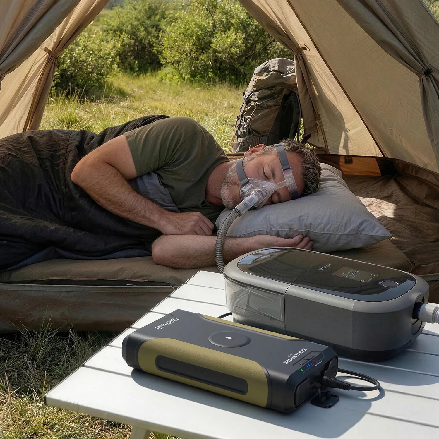 A man sleeps in a tent on a pillow using a CPAP machine powered by a portable battery on a table outside. Camping gear and green vegetation are visible in the background.