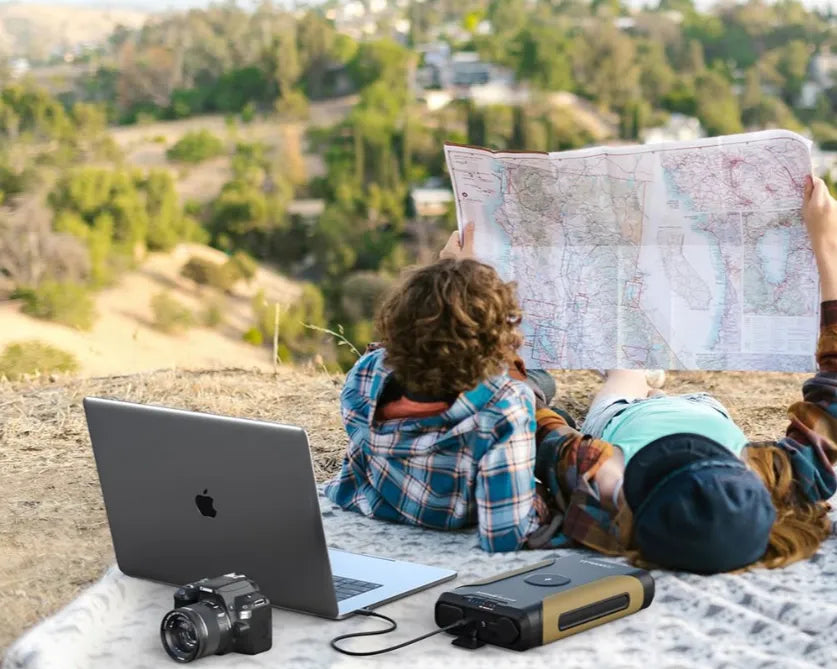 Two people lie on a blanket outdoors, planning their route with a large map. A Solar Generator and binoculars rest nearby, while trees and buildings frame the scene in the background.