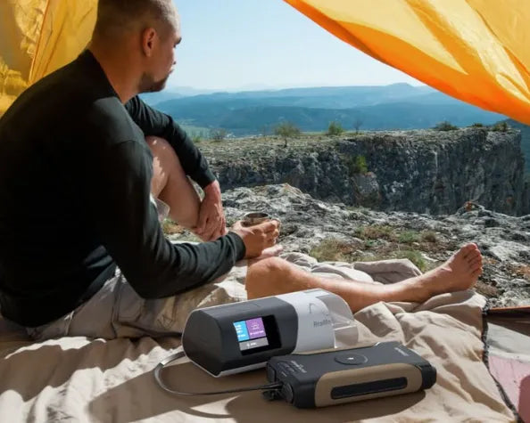 A person sits inside a tent on a rocky surface overlooking a canyon, with a portable CPAP machine and power supply placed in front of them. The tent door is open, revealing the outdoor landscape.