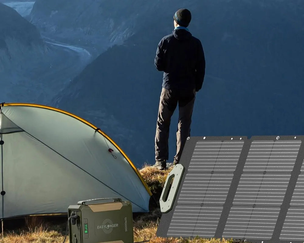 A person stands near a yellow and gray tent on a grassy area overlooking mountains, with a portable power station and foldable solar panel providing camping power nearby.